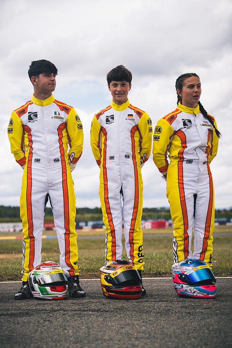 FIA Karting Academy Trophy Senior, Valencia 2026 – Constantin Papst Drei FIA Karting Academy Trophy Fahrer in Rennanzügen stehen nebeneinander auf der Strecke, ihre Helme liegen vor ihnen auf dem Boden; in der Mitte steht Constantin Papst.
