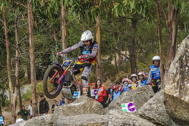 FIM Trial der Nationen (TdN) 2026, Arteixo Ein deutscher Trial-Fahrer überwindet mit seinem Motorrad eine anspruchsvolle Felssektion im Wald, während Teamkollegen und Zuschauer das Geschehen verfolgen.