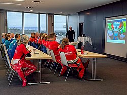 DMSB Medical Days 2026 DMSB Medical Days 2026: Workshop-Teilnehmer in Rennoveralls an Holztischen mit Kaffeetassen; Projektor und Redner zeigen Präsentation "Medical Training".