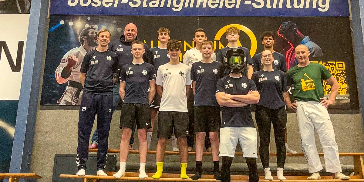 Speedway Motorsport Team Germany 2026 - Saisonauftakt-Fitnesscamp 2026 in Abensberg Gruppenfoto des Motorsport Team Germany Speedway beim Saisonauftakt-Fitnesscamp 2026 in Abensberg; 9 junge Fahrer und 3 Betreuer in Teamkleidung in Turnhalle mit Gymnastikboden stehen auf einer Bank nebeneinander aufgereiht vor Josef-Stanglmeier-Stiftung-Banner.