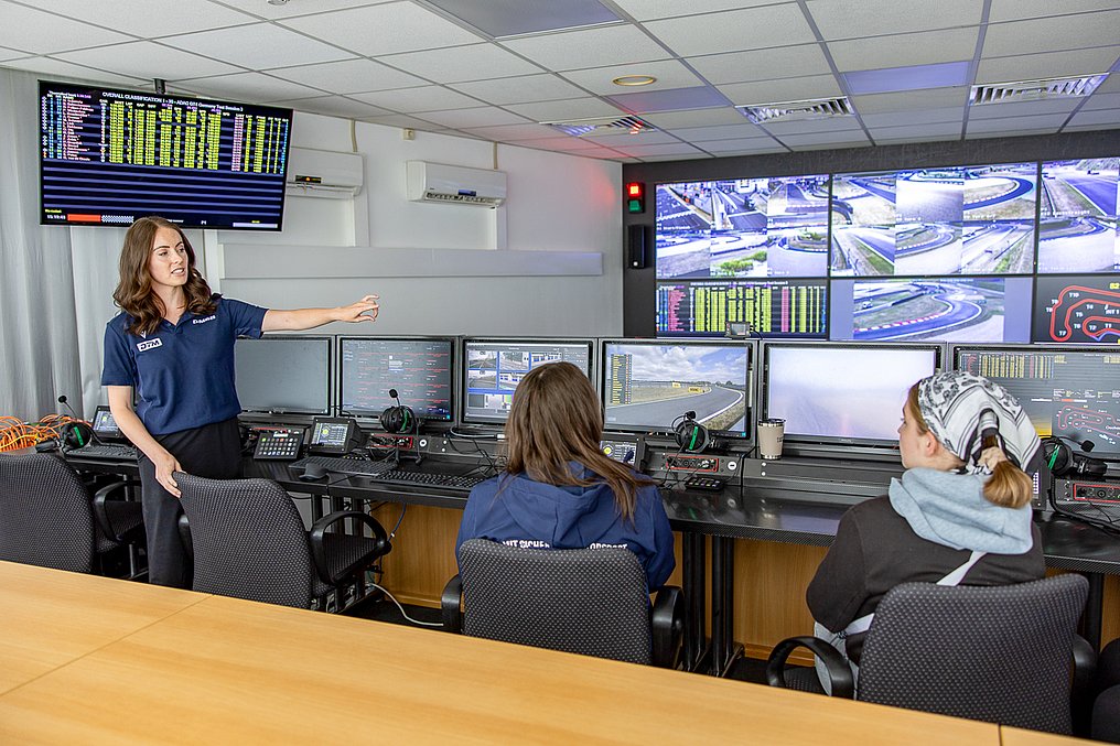 Eine Referentin erklärt im Race-Control-Raum die Monitorwände und die Abläufe der Rennleitung, während Teilnehmerinnen zuhören.