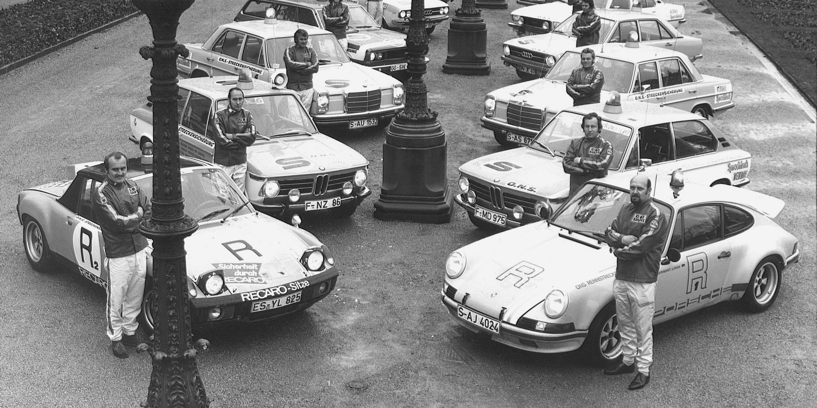 Eine Gruppe von ONS-Staffelaners posiert zum fünfjährigen Jubiläum mit Staffelautos, die in einer Reihe auf einer Straße in der Nähe von verschnörkelten Laternenpfählen stehen.