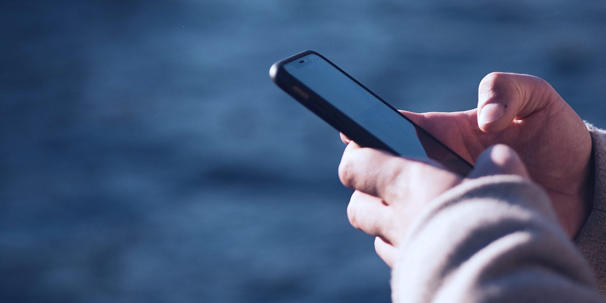 Foto einer Hand, die ein Smartphone hält und auf das beleuchtete Display tippt. Dunkle Dämmerungsstimmung mit blauem Himmel, unscharfem Hintergrund.