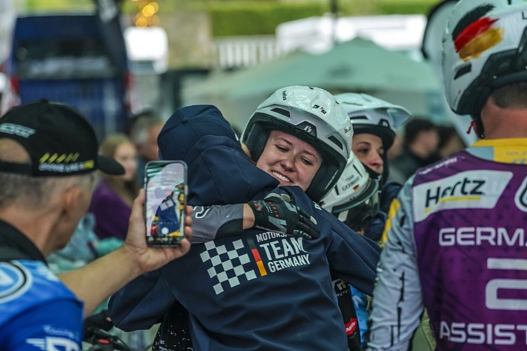 FIM Trial der Nationen (TdN) 2026, Arteixo Eine deutsche Trial-Fahrerin im Helm umarmt ein Teammitglied, während ein Fan den emotionalen Moment mit dem Smartphone festhält.