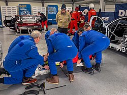 DMSB Medical Days 2026 DMSB Medical Days 2026: Team mit blauen Overalls knien nahe des Bodens, um sich eine Person auf einer Trage zu kümmern; im Hintergrund zwei Rennfahrzeuge, ein Team mit roten Overalls und DMSB-Banner.