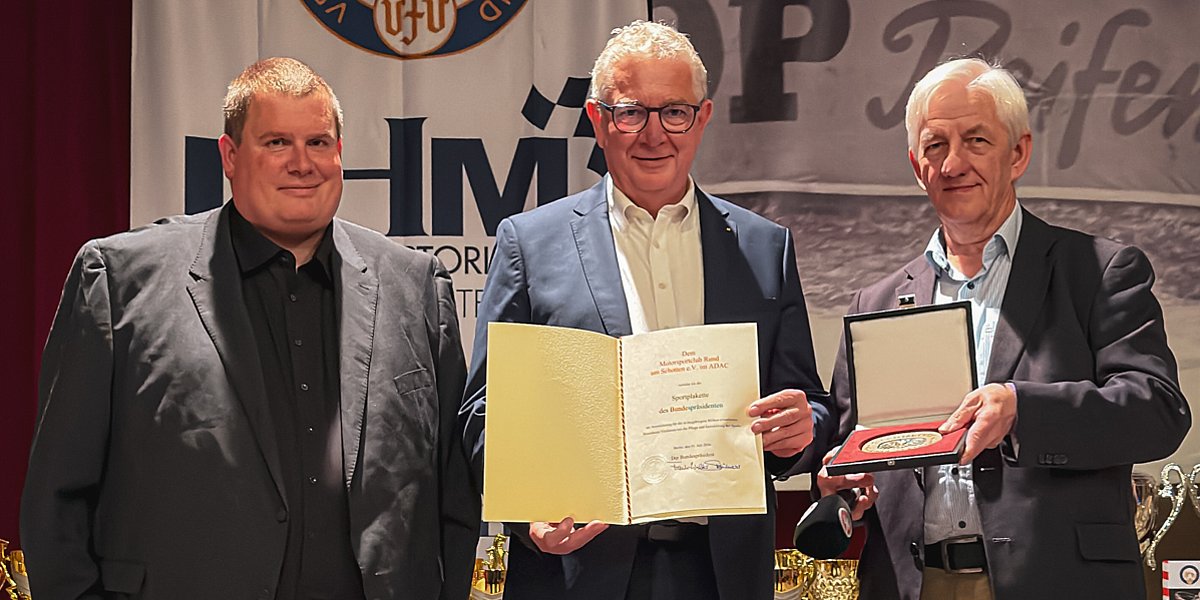 Drei Männer stehen nebeneinander; der Mann in der Mitte hält eine Urkunde in der Hand, und der Mann rechts hält eine geöffnete Prämienbox, in der sich eine Medaille befindet.