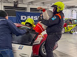 DMSB Medical Days 2026 DMSB Medical Days 2026: Team in dunkelblauen Overalls und gelben Helmen bei Fahrerbergung an weißem Rennwagen; rotes Porsche-Heck sichtbar.