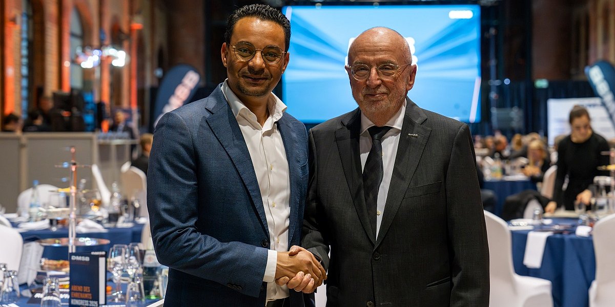 Ein professioneller Handschlag zwischen zwei Männern markiert die Partnerschaft von [BEE]PARTMENT und DMSB beim Kongress 2025. Im Hintergrund eine Event-Halle mit Bühne, Bannern und gedeckten Tischen.