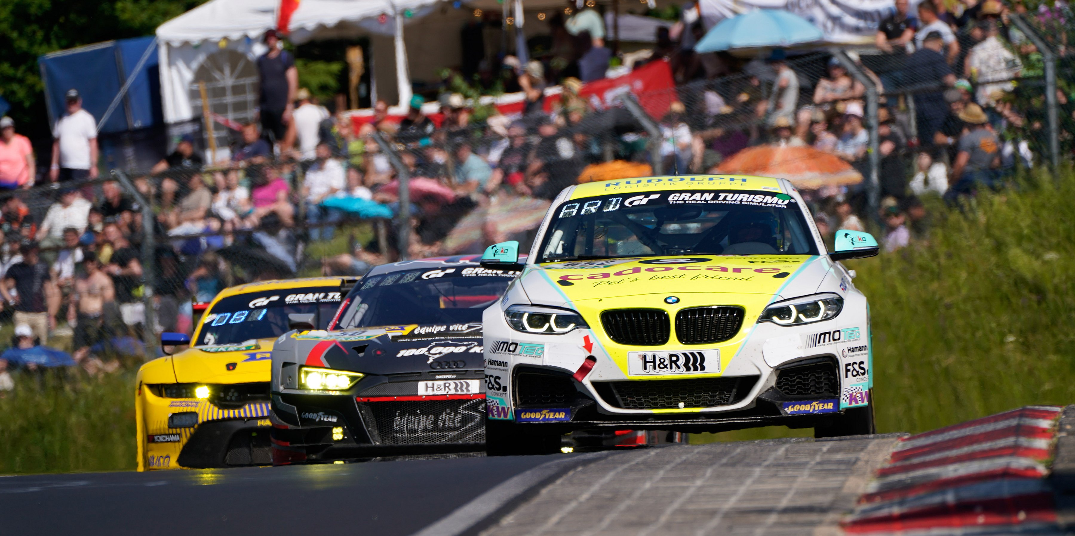 Drei Rennwagen rasen um eine Kurve auf der Rennstrecke, während im Hintergrund eine Menge von Zuschauern zusieht.
