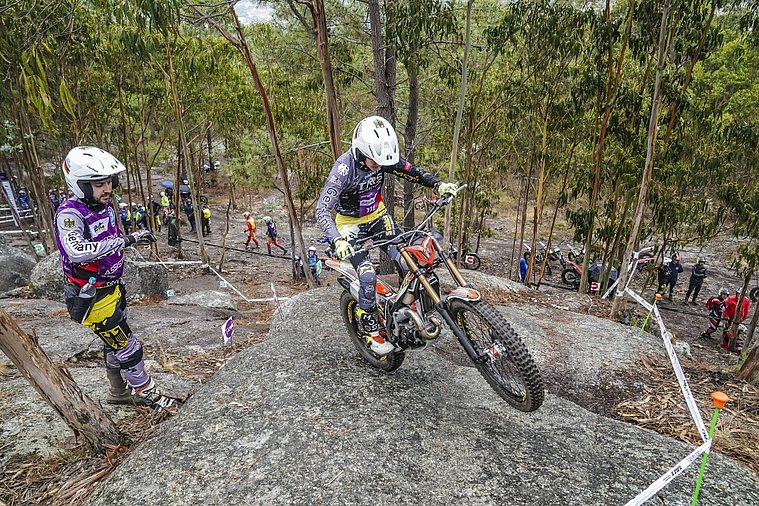 FIM Trial der Nationen (TdN) 2026, Arteixo Eine deutsche Trial-Fahrerin fährt mit ihrem Motorrad über eine felsige Sektion im Wald, während ein Mann in Teamkleidung die Situation beobachtet.