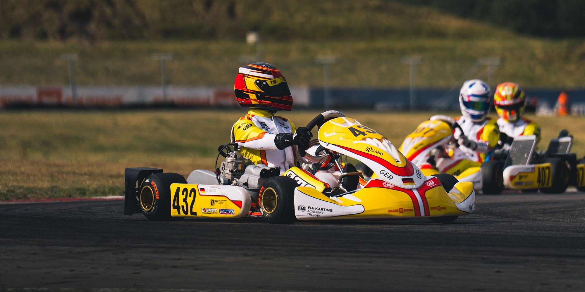 Drei Go-Kart-Fahrer mit Helmen und gelben Anzügen rasen auf einer Rennstrecke. Das führende Kart mit der Nummer 432 wird von zwei anderen dicht gefolgt. Im Hintergrund sind Gras, ein Hügel und ein verschwommener Zaun unter einem teilweise bewölkten Himmel zu sehen.