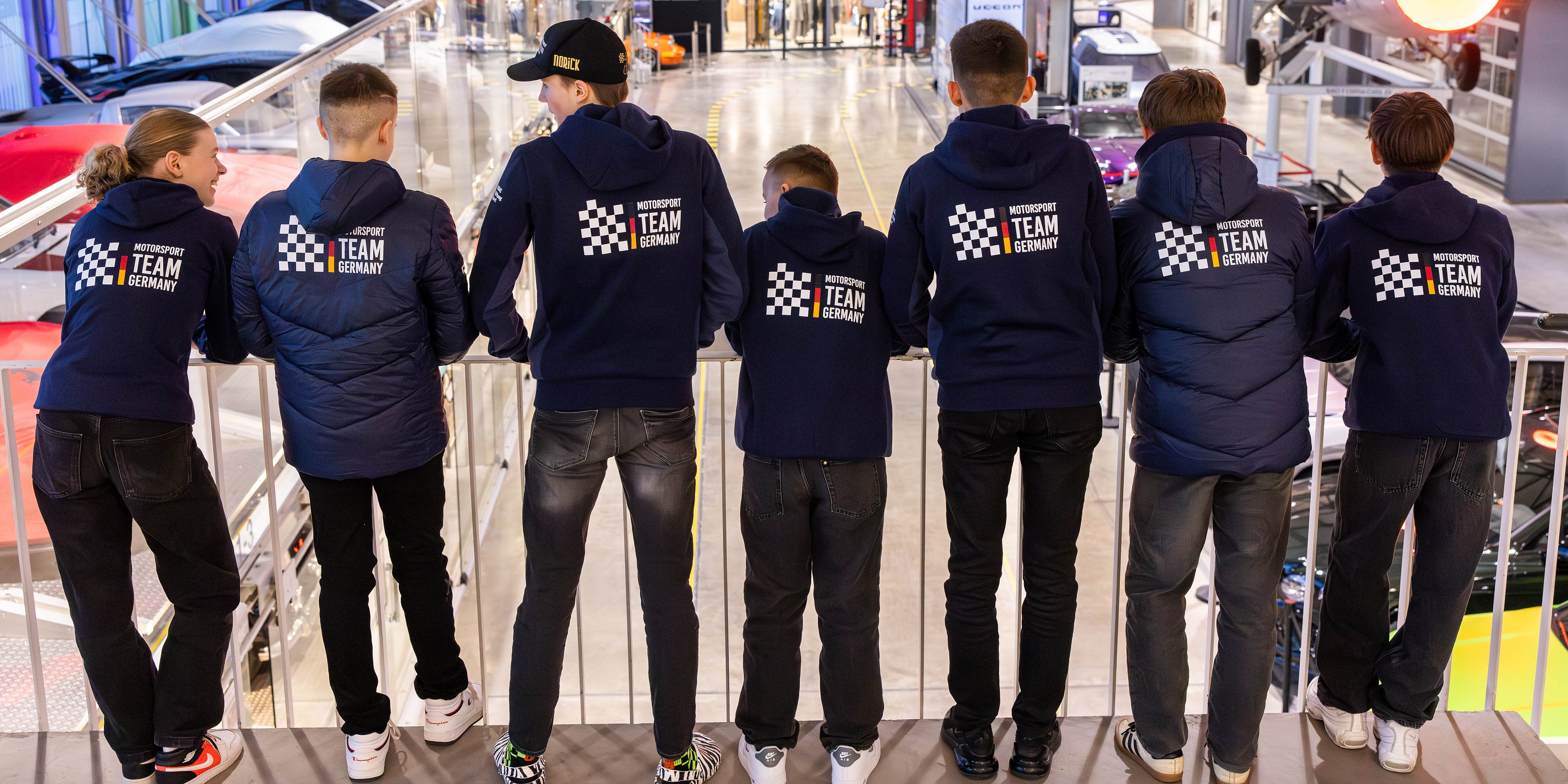 Sieben Personen in passenden Team-Germany-Jacken stehen in einem Innenraum, vor einem modernen Ausstellungsraum mit Autos und Flugzeugen.