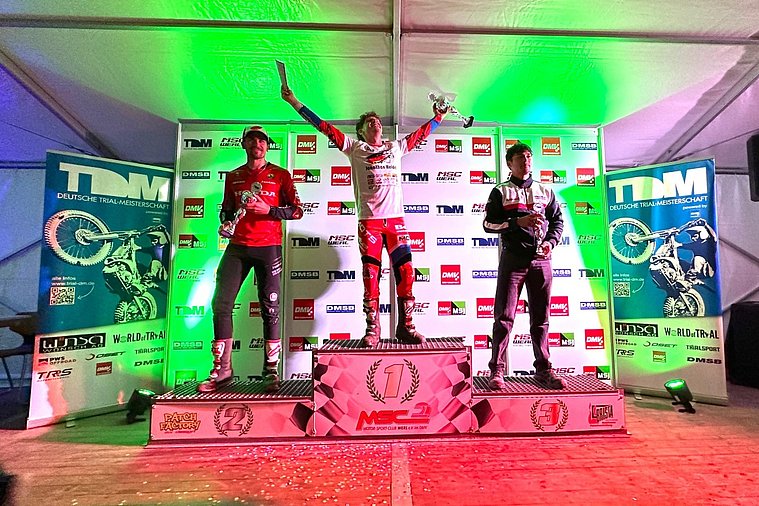 Jonathan Heidel feiert auf dem obersten Podestplatz seinen Sieg beim Finale der Deutschen Trial-Meisterschaft 2026 in Werl. Jonathan Heidel feiert auf dem obersten Podestplatz seinen Sieg beim Finale der Deutschen Trial-Meisterschaft 2026 in Werl. Neben ihm stehen Florian Stephan auf Rang zwei und Rodney Bereiter auf Rang drei mit ihren Pokalen vor der Sponsorentafel.