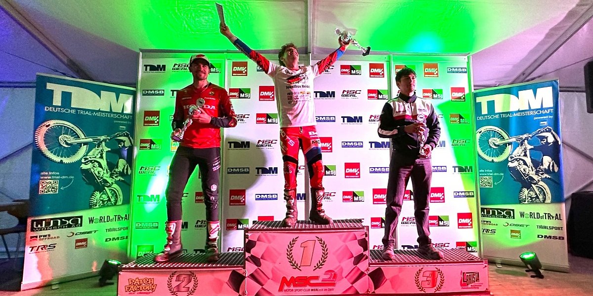 Jonathan Heidel feiert auf dem obersten Podestplatz seinen Sieg beim Finale der Deutschen Trial-Meisterschaft 2026 in Werl. Jonathan Heidel feiert auf dem obersten Podestplatz seinen Sieg beim Finale der Deutschen Trial-Meisterschaft 2026 in Werl. Neben ihm stehen Florian Stephan auf Rang zwei und Rodney Bereiter auf Rang drei mit ihren Pokalen vor der Sponsorentafel.