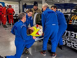 DMSB Medical Days 2026 DMSB Medical Days 2026: Team mit blauen Overalls tragen Frau aus weißem Rennwagen mit offener Tür; im Hintergrund ein Team mit roten Overalls und DMSB-Banner.