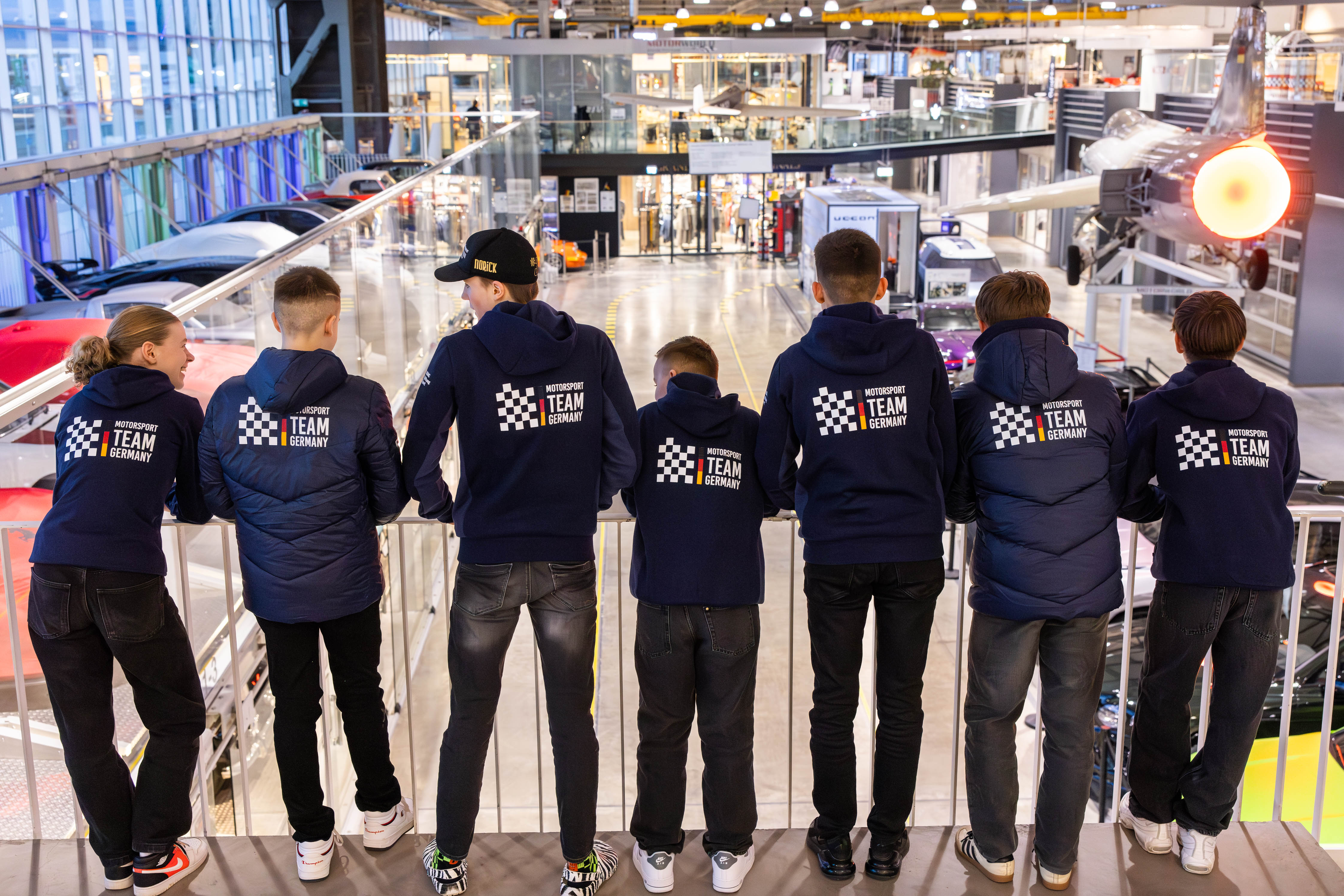 Sieben Personen in passenden Team-Germany-Jacken stehen in einem Innenraum, vor einem modernen Ausstellungsraum mit Autos und Flugzeugen.