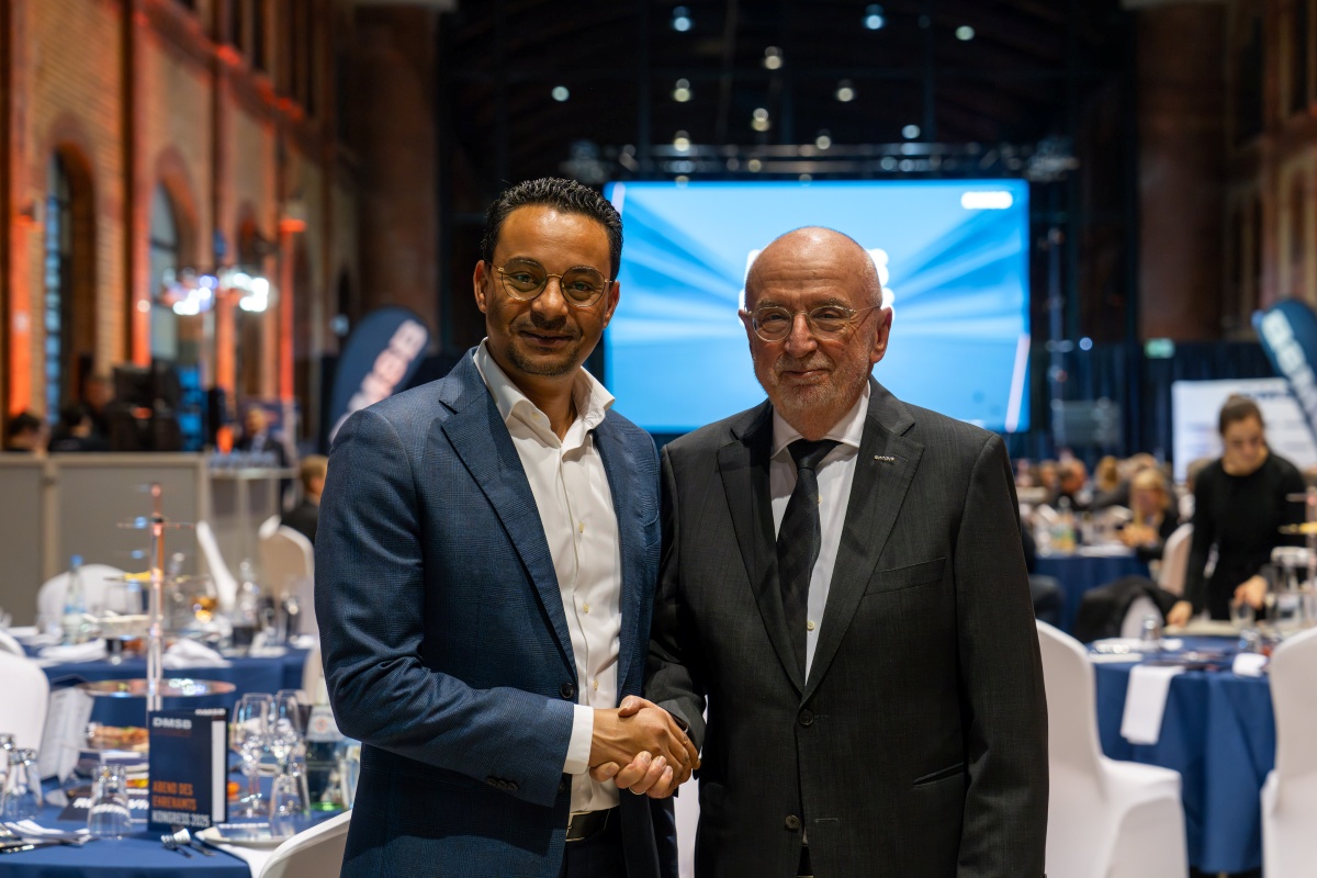 Ein professioneller Handschlag zwischen zwei Männern markiert die Partnerschaft von [BEE]PARTMENT und DMSB beim Kongress 2025. Im Hintergrund eine Event-Halle mit Bühne, Bannern und gedeckten Tischen.