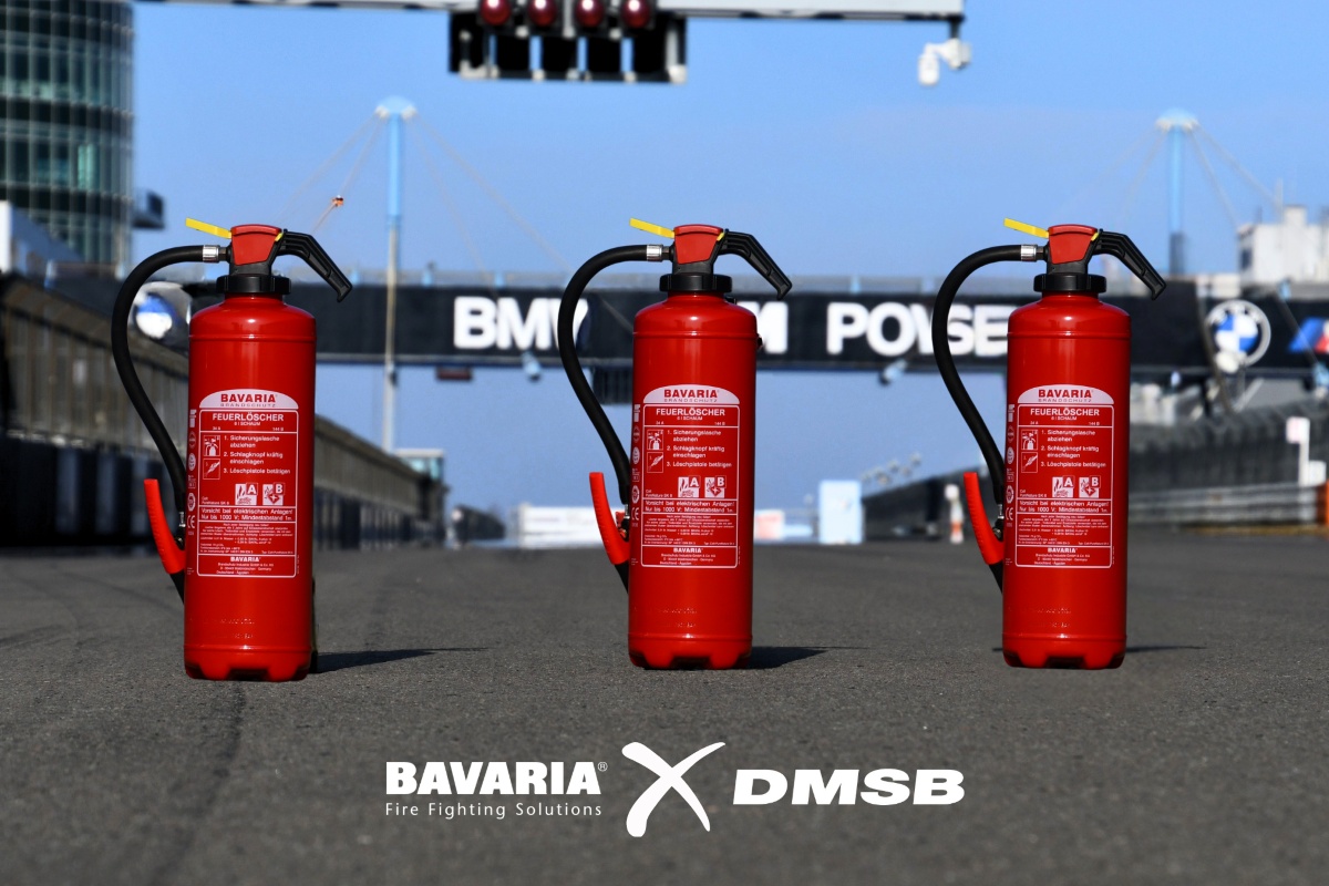 3 rote BAVARIA Fire Fighting Solutions Feuerlöscher stehen auf einer asphaltierten Rennstrecke bei blauem Himmel. Unten die Logos von BAVARIA x DMSB in weiß.