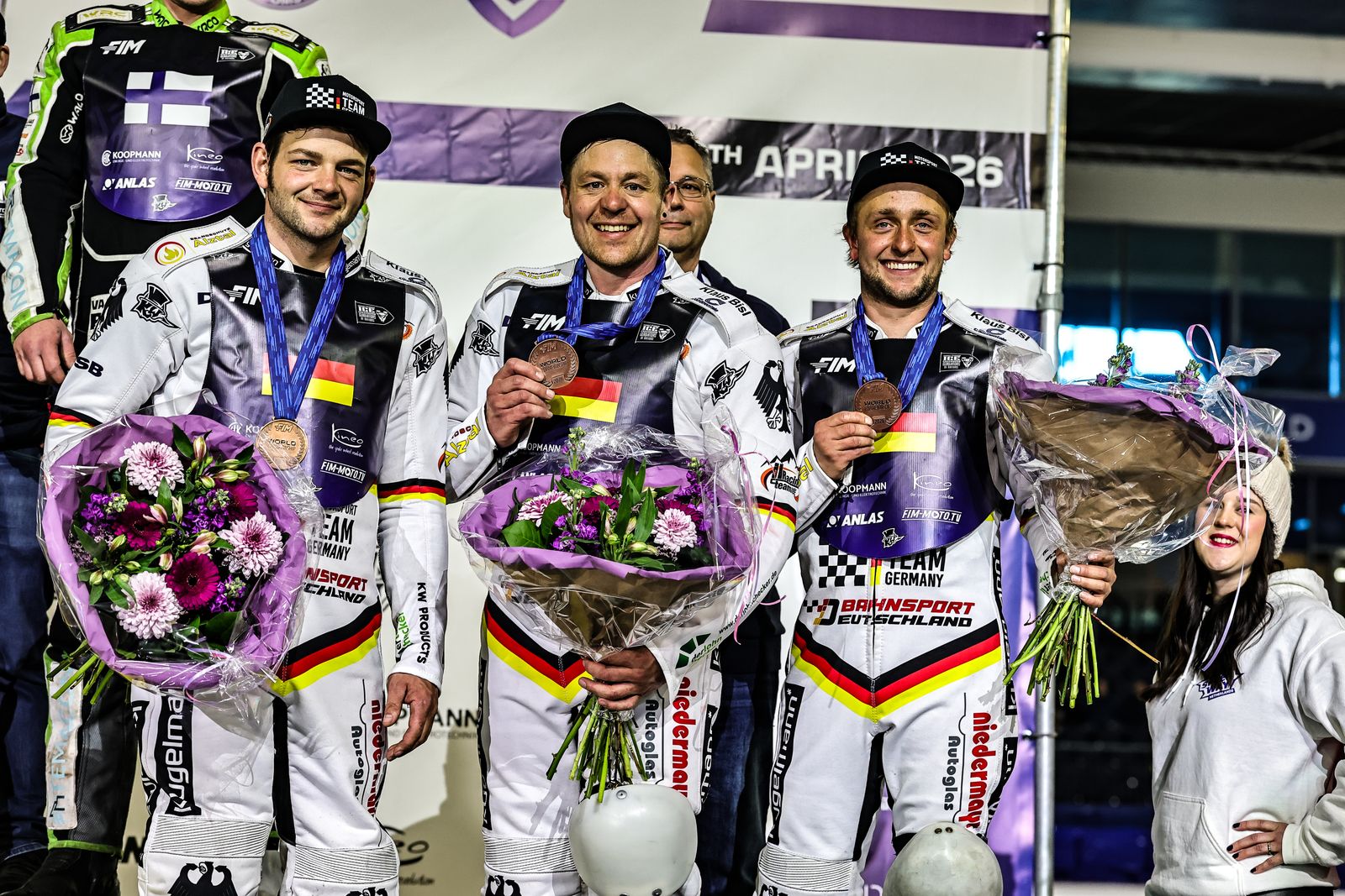 Drei lächelnde Motorradrennfahrer in weißen Uniformen halten Medaillen und Blumensträuße in der Hand und stehen auf einem Podium in einer Halle.