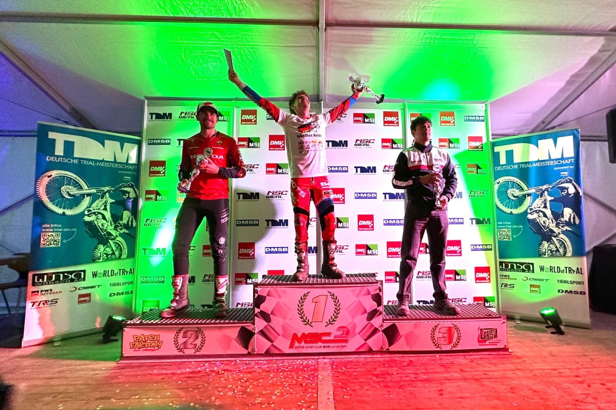 Jonathan Heidel feiert auf dem obersten Podestplatz seinen Sieg beim Finale der Deutschen Trial-Meisterschaft 2026 in Werl. Neben ihm stehen Florian Stephan auf Rang zwei und Rodney Bereiter auf Rang drei mit ihren Pokalen vor der Sponsorentafel.