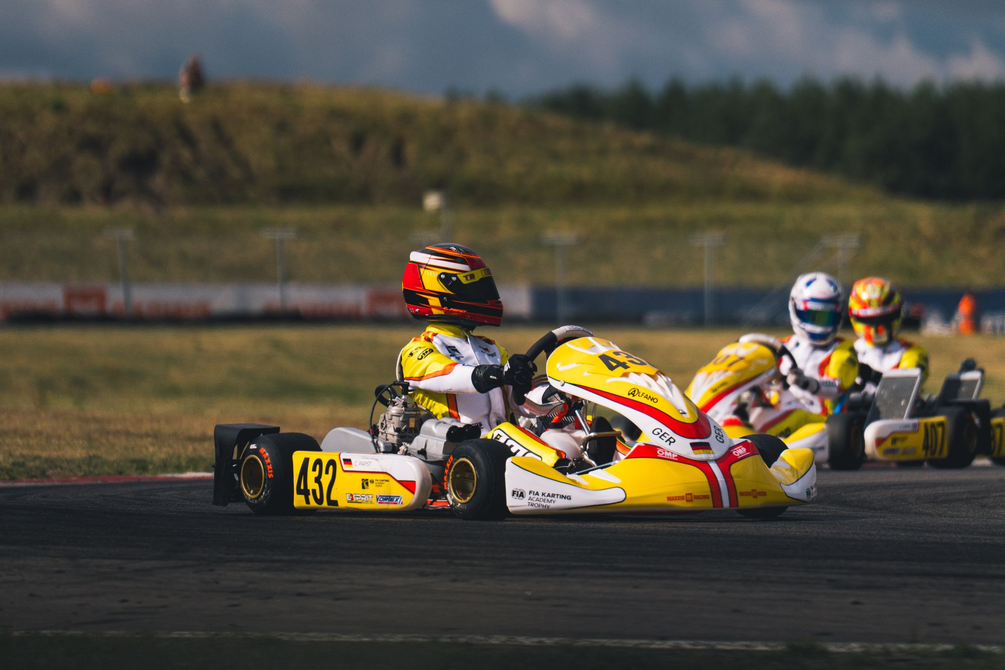 Drei Go-Kart-Fahrer mit Helmen und gelben Anzügen rasen auf einer Rennstrecke. Das führende Kart mit der Nummer 432 wird von zwei anderen dicht gefolgt. Im Hintergrund sind Gras, ein Hügel und ein verschwommener Zaun unter einem teilweise bewölkten Himmel zu sehen.